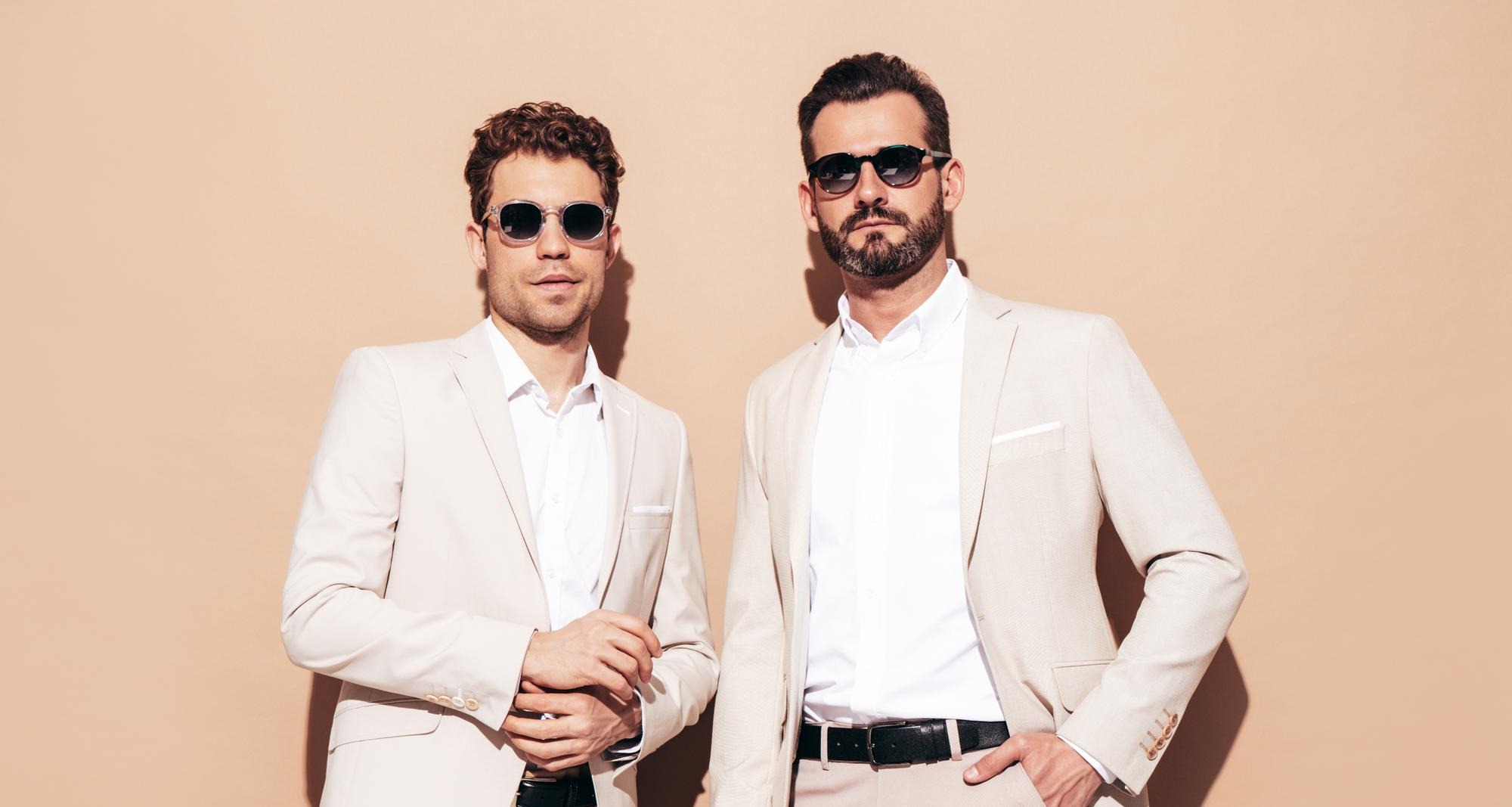 Two stylish men in light beige suits wearing sunglasses, posing against a tan backdrop.